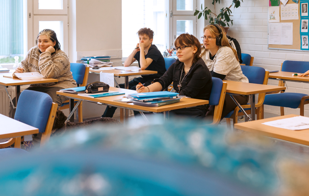 I framgrunden ser man suddigt övresidan av en stor jordglob. I bakgrunden sitter fyra studerande i klass vid sina pulpeter och lyssnar på läraren.