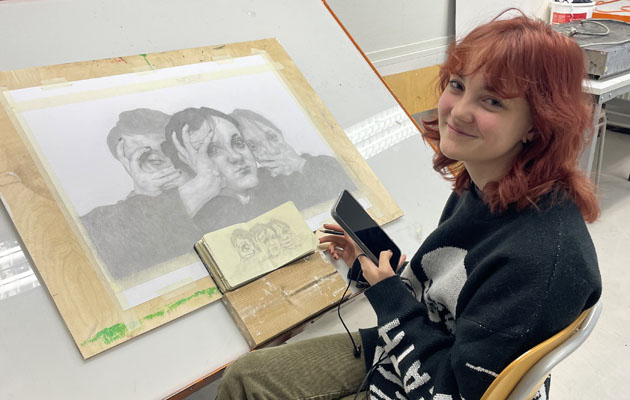 Ung studerande sitter vid sin blyertsteckning och tittar glatt in i kameran.