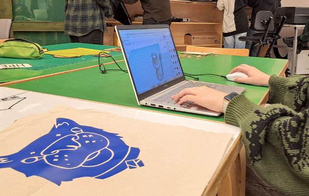 I förgrunden ser man en vit tygkass med ett blått tryck som påminner om ett kattansikte. Bakom ser man händerna på någon som arbetar vid en laptop på vars skärm man ser en schackpjäs design.