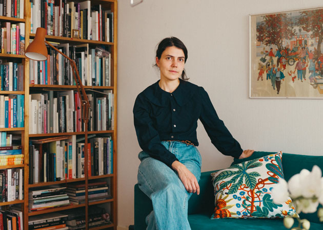 Ida Taavitsainen sittandes på en grön soffas armstöd. I soffan finns en blommig kudde och i bakgrunden en bokhylla i träd fylld av böcker.