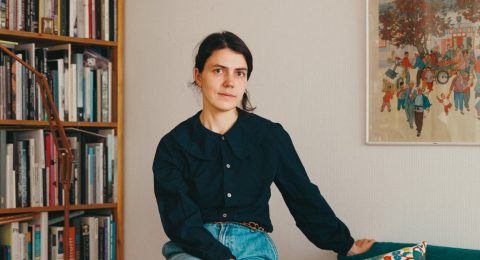 Ida Taavitsainen sittandes på en grön soffas armstöd. I soffan finns en blommig kudde och i bakgrunden en bokhylla i träd fylld av böcker.