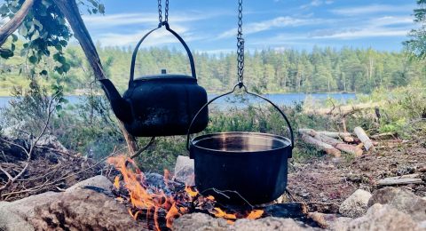 Matlagning utomhus. I framgrunden hänger en stor kaffepanna och en gryta över öppen eld. I bakgrunden ser man en sjö, granskog och blå himmel.