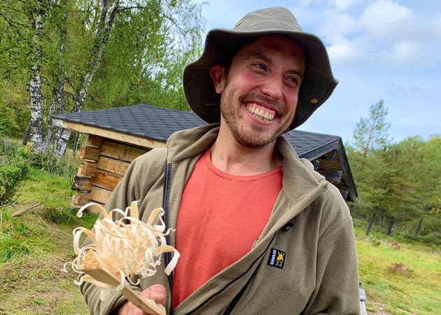 Porträtt av Fred Södergran som ler stort och har en mörkgrön bushhatt på huvudet. I bakgrunden skymtar en liten stockstuga.