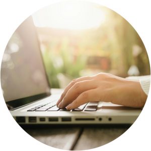 Närbild av händer på ett tangentbord. Laptopen står på ett trädbord utomhus. I bakgrunden ser man starkt solljus.