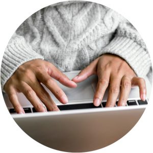 Närbild av händer som skriver på laptop. Personen har en vit stickad tröja på sig.